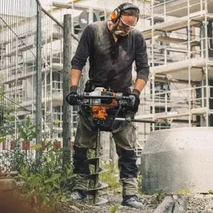 a man with a large moustache, wearing ear defenders, and using the STIHL BT 131 Petrol Earth Auger to drill a hole in the ground on a building site