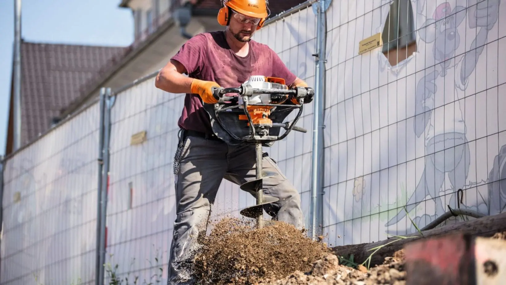 a man wearing a hard hat, using the STIHL BT 131 Petrol Earth Auger to dig a hole in the ground on a building site, next to a housing development