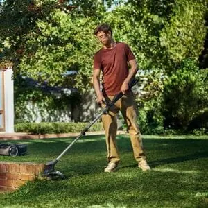 a man using the Husqvarna Aspire T28-P4A Battery Grass Trimmer at the edge of a lawn, with a wall, robot mower, and lots of trees in the background
