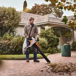 a man on a driveway outside a house, using the SHA 56 Cordless Vacuum Shredder to collect a pile of dry leaves
