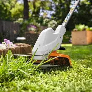 a close up of the STIHL FSA 30 Cordless Grass Trimmer cutting long grass in a garden, with planters, fencing and trees in the background