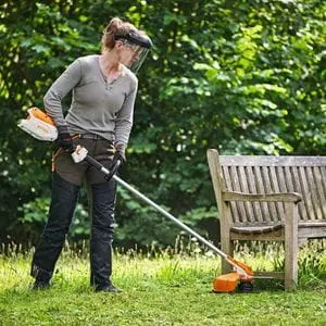 a woman wearing a face shield, using the STIHL FSA 86 R Cordless Brushcutter to trim long grass underneath a wooden bench