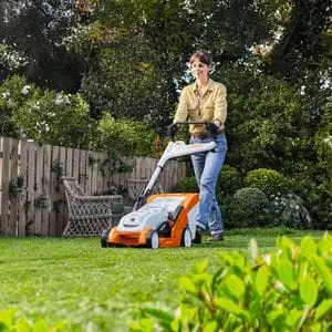 a woman mowing a lawn using the STIHL RMA 235, with trees, shrubs, a fence and wooden bench in the background