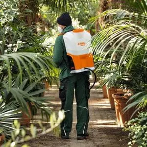 a man stood on a path between large numbers of potted plants, operating the STIHL SGA 85 Cordless Sprayer, which is powered by a backpack battery