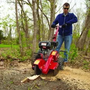 a man wearing sunglasses, using the Toro SGR-13 Petrol Stump Grinder to grind down a tree stump, with other trees in the background
