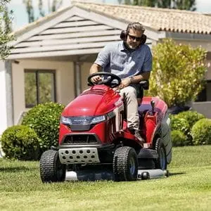 a man wearing sunglasses and ear defenders, mowing a lawn using the Honda HF 2625 Premium Petrol Lawn Tractor, with a bungalow, trees and shrubs in the background