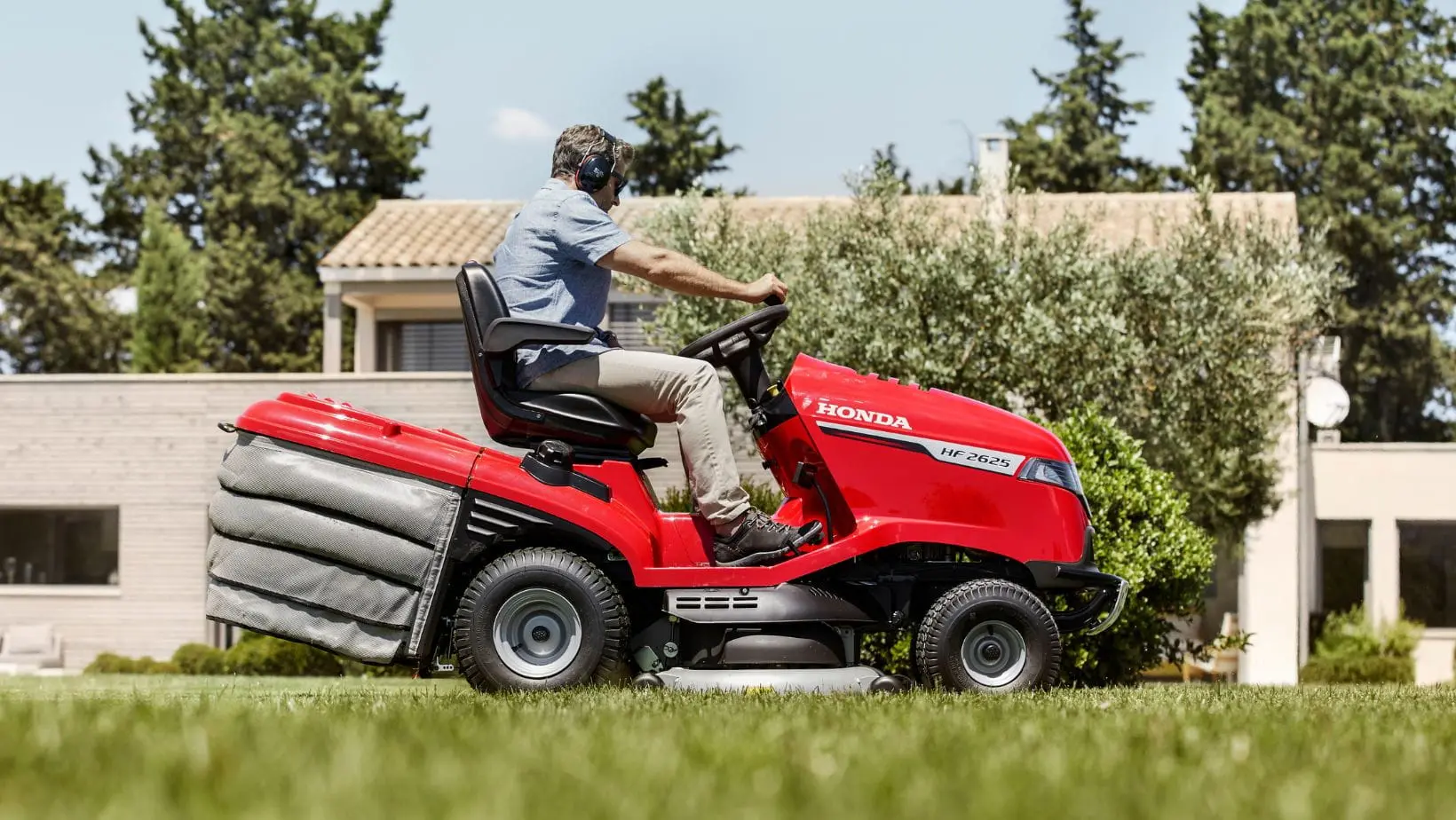 a side shot of a man driving the Honda HF 2625 Premium Petrol Lawn Tractor across a lawn, with a house and trees in the background