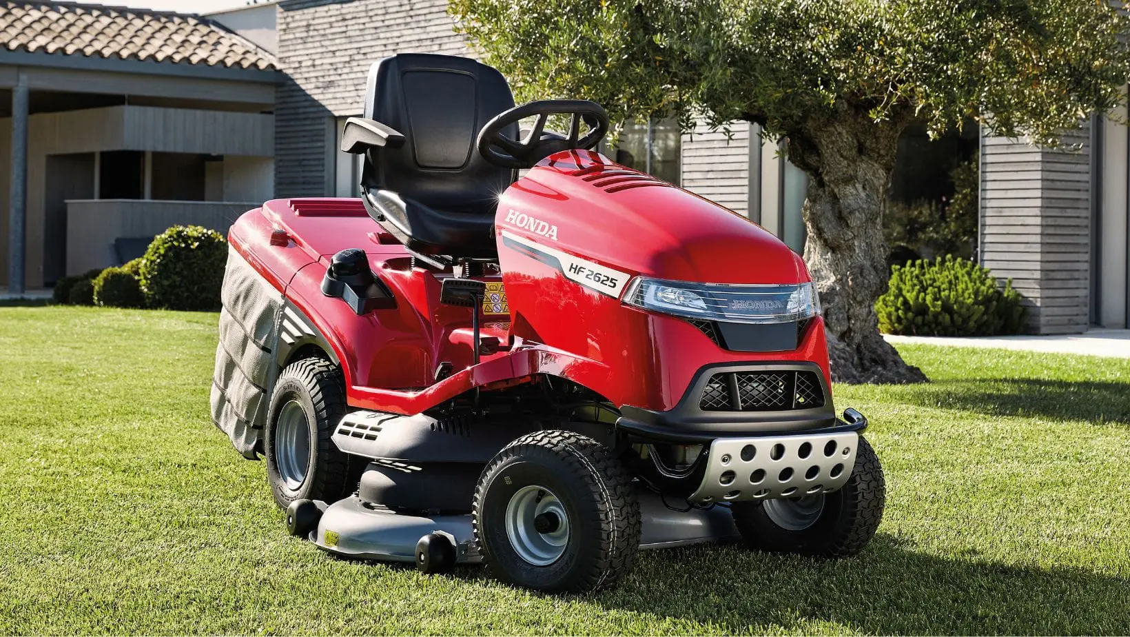the Honda HF2625 Premium Petrol Lawn Tractor parked on a lawn, with a tree and large house in the background