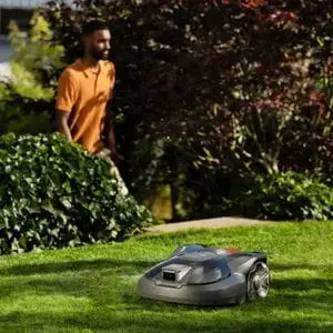 a Husqvarna Automower® 308V Robotic Mower on a lawn, with a man dressed in an orange polo shirt walking up some steps onto the lawn behind it