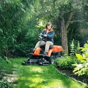 a woman steering the Husqvarna R 112C Petrol Compact Rider around a flowerbed on an intricate lawn