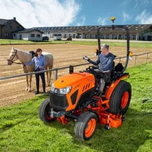 a man sat on a Kubota LX compact tractor, speaking to a woman who is stood on an equestrian track holding a horse's bridle
