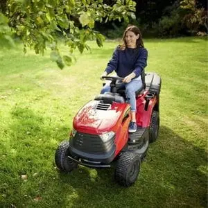 a woman with long, dark hair, driving the Mountfield 1330M Petrol Garden Tractor across a lawn and under a tree canopy