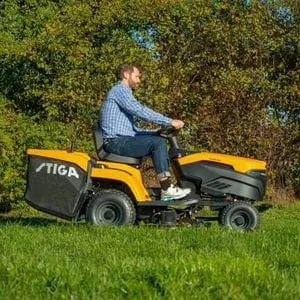 a man driving a STIGA Estate 384e Battery Garden Tractor across an area of grass, with trees in the background