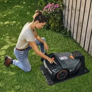 a woman kneeling on one knee and manually starting a STIHL iMow Robotic Mower, which is sat in its charging station near a fence