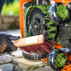 someone wearing black, STIHL-branded gloves, using a brush to clean the underside of a STIHL iMow Robotic Mower