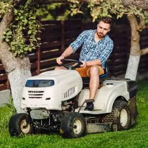 a man wearing shorts and a shirt, using a white riding mower to cut a lawn, with 2 trees and a row of slatted fence panels in the background