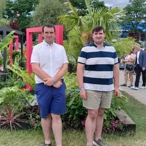 2 men dressed in shorts, stood in front of Dakic Garden Design's The Heart of the Jungle show garden, with square, pink arches and extensive foliage prominent in the background