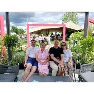 2 men and 2 women, sat on a bench, underneath a grey gazebo, with square, pink arches and green foliage prominent in the background - part of Dakic Garden Design's The Heart of the Jungle garden