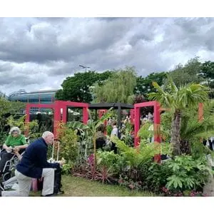 a man knelt on one knee and a woman in a wheelchair, admiring Dakic Garden Design's The Heart of the Jungle show garden, featuring square, pink arches, a grey gazebo, plus extensive trees and plants