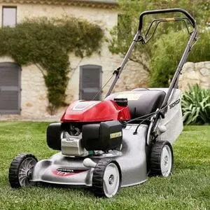 the Honda Izy HRG 466 SKEH Petrol Lawn Mower on a lawn, next a large house made from lightly-coloured brick