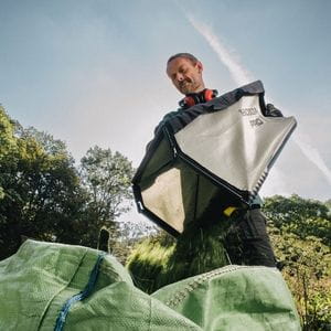 a man emptying clippings from a lawnmower's grass bag into a large, green sack