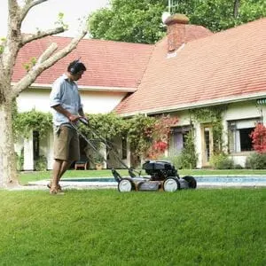 a man wearing ear defenders, a shirt and shorts, using a Husqvarna mulching mower to cut a lawn, next to a small outdoor swimming pool and large bungalow