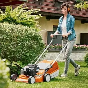 a woman wearing ear defenders, using a STIHL mower to cut grass near a hedge, in front of a house