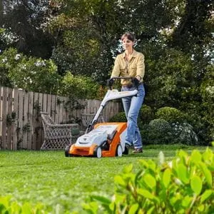a woman mowing a lawn using a STIHL mower, with trees, shrubs, fencing and a wooden bench in the background