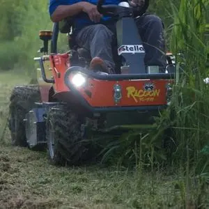 the Stella Racoon 110 Ride-on Flail Mower, with headlights on, cutting long grass on the side of a dirt path