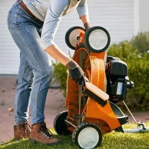 a woman brushing clean the underside of an orange STIHL lawnmower