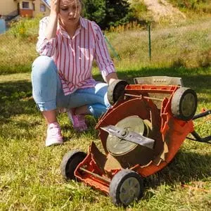 a young woman looking frustrated, crouched down next to an old, orange, upturned lawnmower