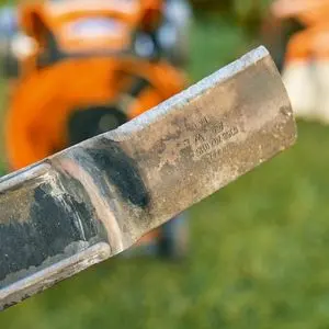 a close up of a metal lawnmower blade, with an orange lawnmower in the background