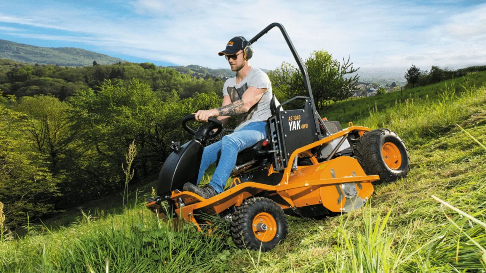 a man cutting long grass on a very steep hill using the AS-Motor AS 1040 YAK 4WD XL Ride-on Flail Mower, with panoramic views of the countryside in the background