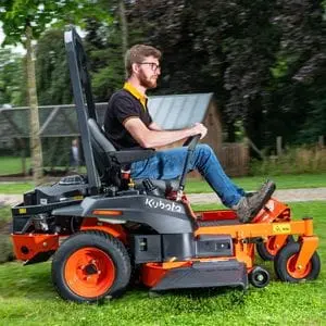 a man cutting grass using the Kubota Z1-421 Petrol Zero-Turn Mower, with a path, greenhouse and trees in the background