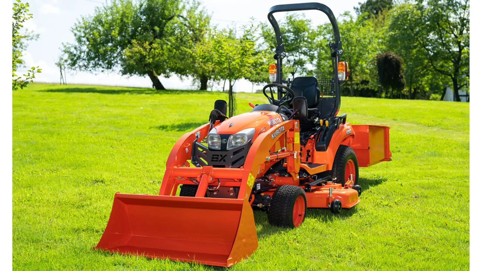 a Kubota compact tractor fitted with front loader, mowing deck and rear-mounted container, parked in a field