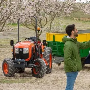 a man stood in front of a Kubota small tractor, which is fitted with a trailer and parked beneath a tree