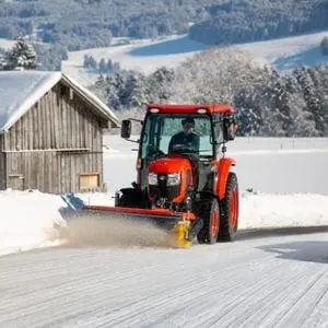 a Kubota compact tractor fitted with a snow plough attachment, clearing snow from a country road 