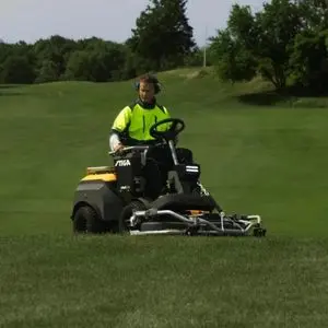 a man wearing a high-visibility jacket and ear defenders, cutting grass in the middle of a golf course using the STIGA ePark Pro Battery Front Mower