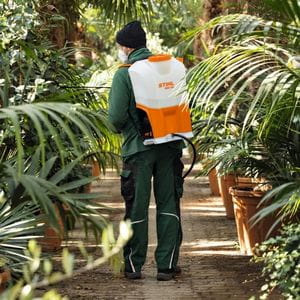a man wearing green overalls, a hat and face mask, using the STIHL SGA 85 Cordless Sprayer to apply treatment to tropical plants
