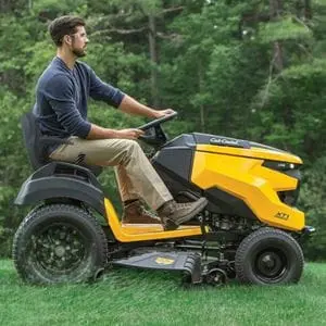 a man cutting grass next to a wood, using a yellow Cub Cadet lawn tractor