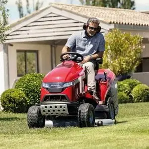 a man wearing ear defenders, using the Honda HF 2625 HTE Premium Petrol Lawn Tractor to cut a lawn outside a bungalow
