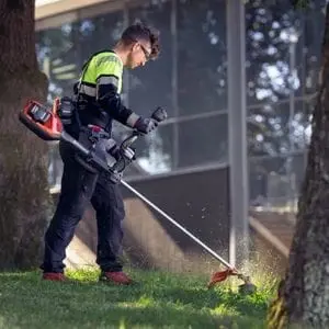 a man using the Husqvarna 525iRXT Brushcutter to cut grass in between trees, near a large building