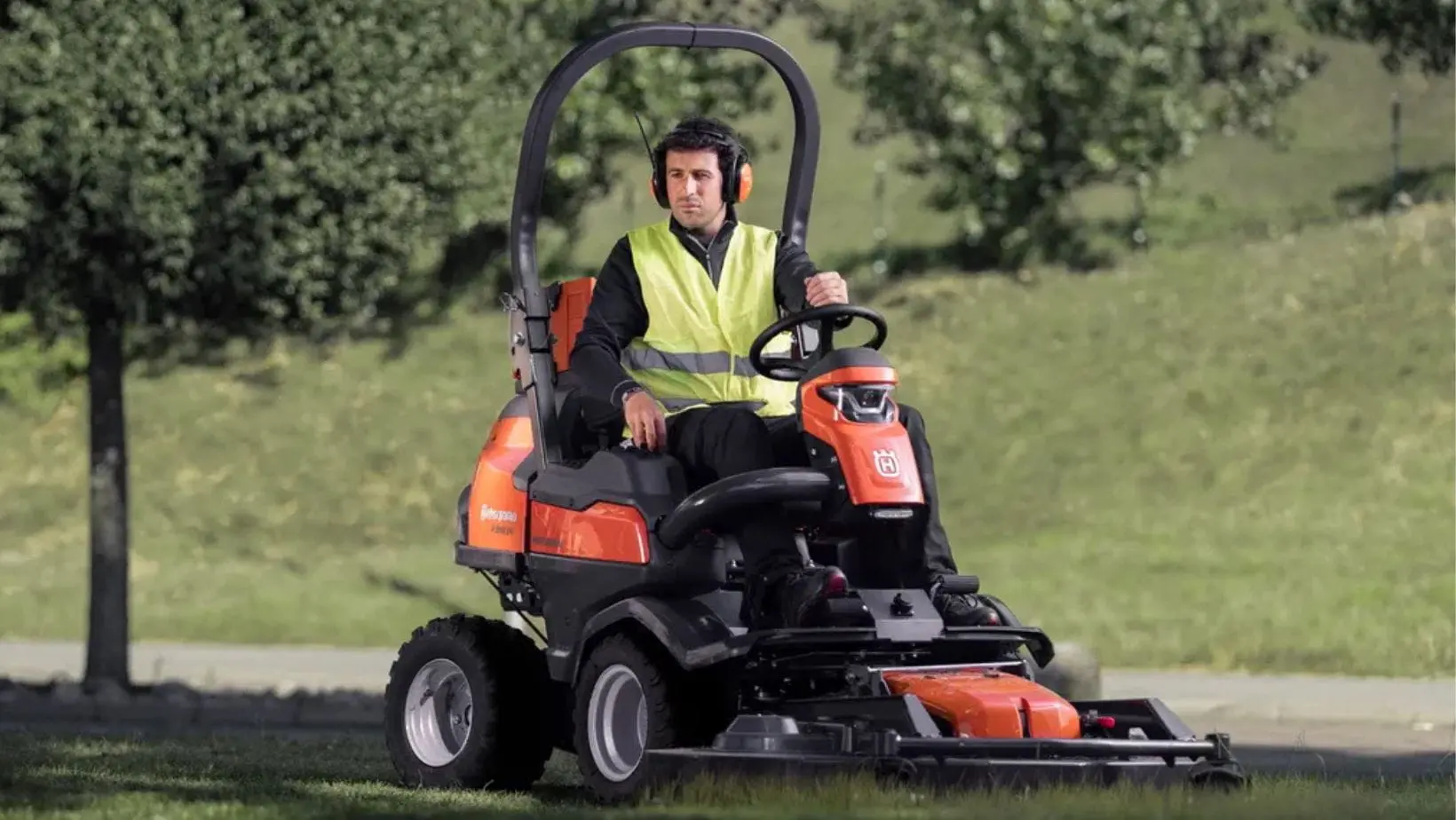 a man wearing a yellow high-visibility jacket and ear defenders, navigating the Husqvarna P 524X Out Front Mower past a tree