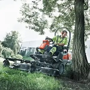a man using the Husqvarna P 525DX Out Front Mower to cut grass around a tree, outside a large building