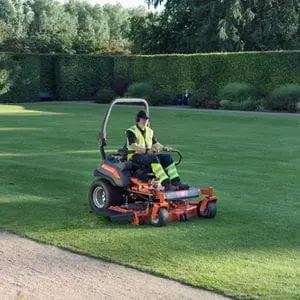 a man in a high-visibility jacket, mowing a large ornamental lawn using a Husqvarna zero turn mower