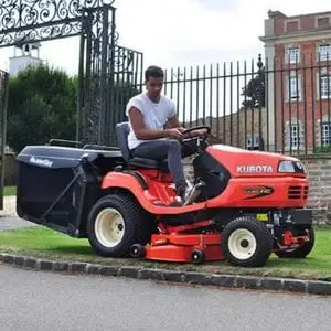a man using the Kubota G21E Ride-On Mower to cut a small patch of grass outside the gate that leads to a large building