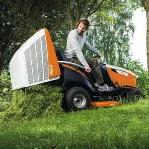 a man sat in the driver's seat of the STIHL RT 6112 Petrol Ride-on Lawn Mower, depositing grass from the low-dump collector near some trees, at the edge of a field