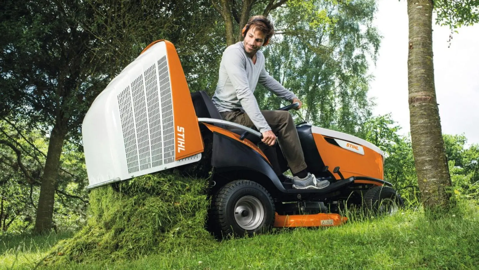 a man sat on the STIHL RT 6112 Petrol Ride-On Lawn Mower, emptying grass from its collector next to some trees