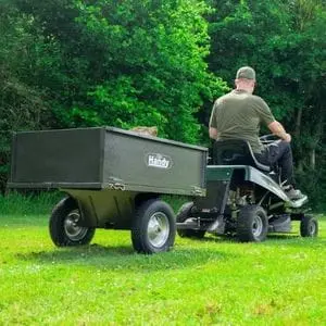 a man sat on a riding mower towing the Handy THGT500 Towed Steel Tipping Trailer across a lawn