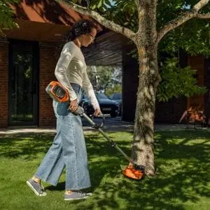 a woman using the Husqvarna 215iL Grass Trimmer to cut a lawn around a tree, outside a house